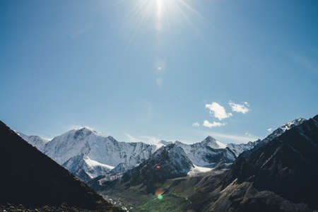Alpine landscape with high snowy mountain with peaked top under cirrus clouds in sun with lens flare in sky. Big snow covered mountains in sunshine. Black rocks and white-snow pointy peak in sunlight.の写真素材