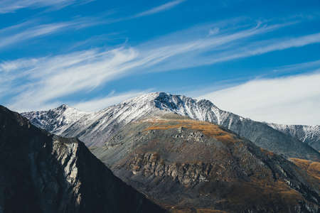 Colorful alpine landscape with great mountain in autumn colors with snow on peak in sunshine under cirrus clouds in blue sky. Picturesque autumn scenery with sunlit rocks and snow-covered mountain topの写真素材