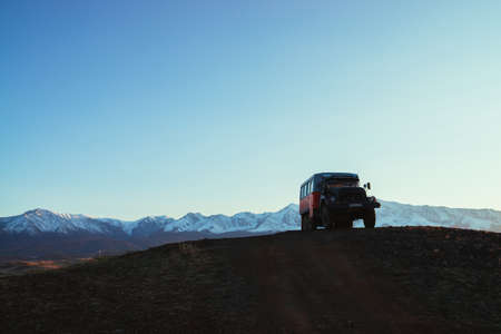 Truck on hill with view to great snow mountain range in sunlight. Sunset landscape with truck on background of snowy mountains in sunshine under blue sky. Russia, Altai Republic, 20 September, 2020.のeditorial素材