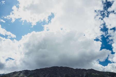 Awesome scenery with shadow of big cloud on high mountain top. Huge white cloud in blue sky over giant black rocky mountain peak. Scenic landscape with high dark rock in shadow of cloud in sunny day.の写真素材