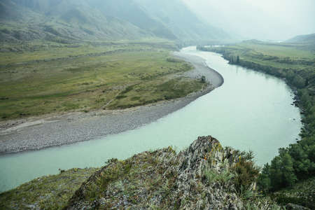 Rock with grasses and lichens with view to big mountain river in mist. Beautiful mountain landscape with big rock with flora on background of wide mountain river in rainy weather. Atmospheric scenery.の写真素材