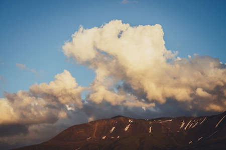 Atmospheric mountain scenery with yellow clouds in blue sky. Scenic landscape with illuminating sunset clouds above mountains with snow. Beautiful sunrise in mountains. Snow on rocks in sunlight.の写真素材