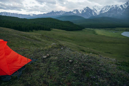 Atmospheric alpine landscape with orange tent on background of lake and big snowy mountains in overcast weather. Awesome green scenery with orange tent with view of great mountains under cloudy sky.の写真素材