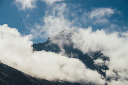 Minimalist view of snow-capped mountain silhouette above thick clouds. Scenic mountain landscape with white-snow sharp peak among dense low clouds in blue sky. Wonderful scenery with snowy pinnacle.の写真素材