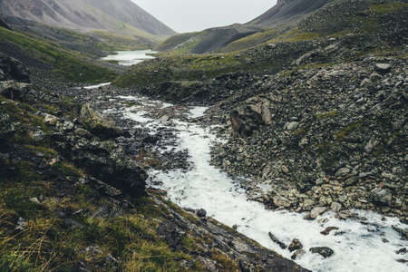 Atmospheric landscape with mountain lake and mountain creek among moraines in rainy weather. Bleak overcast scenery with milky river and lake among rocks. Gloomy view to milk mountain river and lake.の写真素材