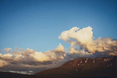 Atmospheric mountain scenery with yellow clouds in blue sky. Scenic landscape with illuminating sunset clouds above mountains with snow. Beautiful sunrise in mountains. Snow on rocks in sunlight.の写真素材