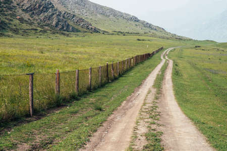 Beautiful green mountain landscape with long dirt road along fence and big mountains in fog. Atmospheric foggy mountain scenery with dirt road among rocks and big mountains. Length road in countrysideの写真素材