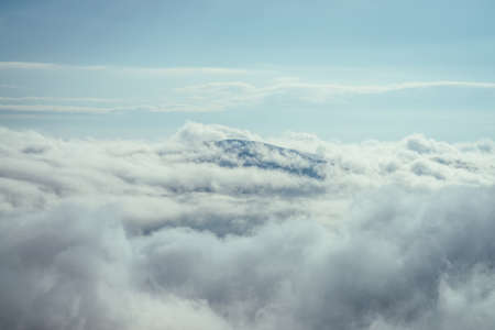 Wonderful minimalist landscape with mountain top above dense low clouds. Mountain vertex floats in thick clouds. Scenic minimalism with mountain peak above cloudy sky. Beautiful summit in cloudiness.の写真素材
