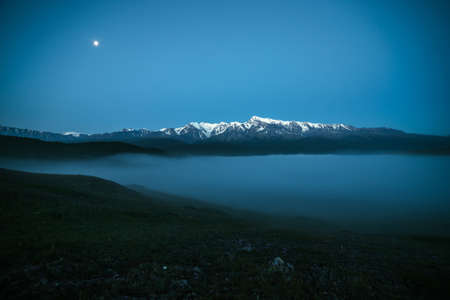 Atmospheric mountains landscape with dense fog and great snow mountain top under twilight sky....
