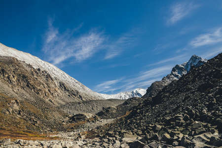 Awesome alpine landscape with dark rockies and high snow-covered mountains in sunshine under blue sky with cirrus clouds. Beautiful highland scenery with sharp black rocks and mountain top with snow.の写真素材