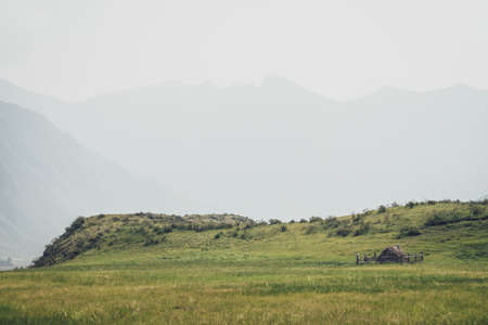 Beautiful foggy mountain landscape with scarecrow on wooden fence around haystack near hills on background of mountains silhouettes in fog. Stack of hay is surrounded by wood fence in vintage tones.の写真素材