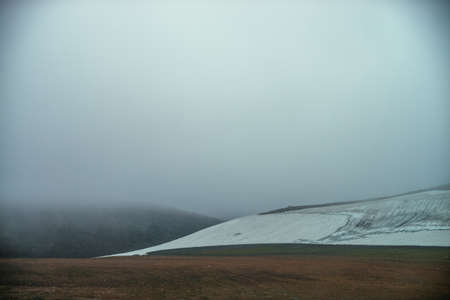Atmospheric minimalist mountain landscape with small glacier on rocky hill slope inside low cloud. Mountainside with small glacier in dense fog. High mountain valley in low cloud. Poor visibility.の写真素材