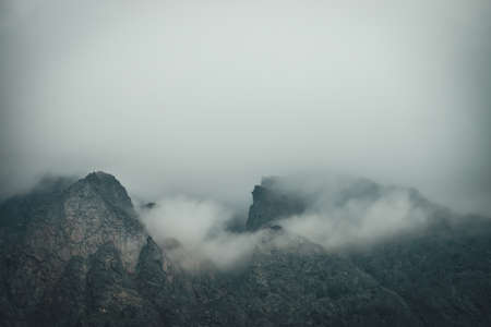 Dark atmospheric mountain landscape with sharp rocks in low clouds. Dark rocks in gray overcast weather. Gloomy mountain scenery with gray low clouds on rough crags. Minimalism with rocky mountains.の写真素材