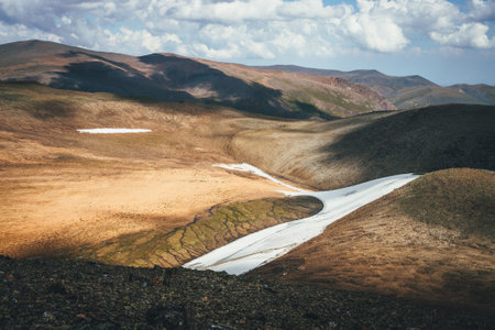 Sunny mountain desert relief with long glacier on slope in sunlight under cloudy sky. Sunny scenic highlands landscape with long small glacier on mountainside and melt water streams high in mountains.の写真素材