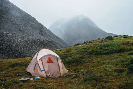 Scenic alpine landscape with tent on green hill among rocks. Beautiful snow-capped mountains in low clouds. Atmospheric scenery with tent in highlands in overcast weather. High mountains covered snow.の写真素材