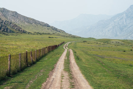 Beautiful green mountain landscape with long dirt road along fence and big mountains in fog. Atmospheric foggy mountain scenery with dirt road among rocks and big mountains. Length road in countrysideの写真素材