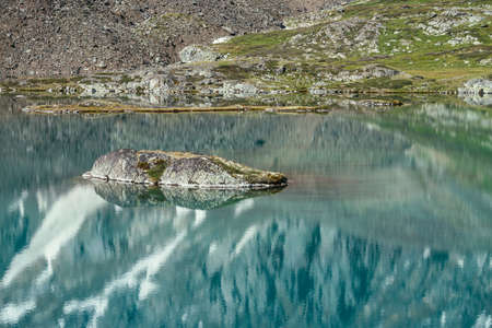 Rock in turquoise mountain lake. Snowy mountain reflected in azure clear water of glacial lake. Beautiful sunny background with snow-white glacier reflection in green water surface of mountain lake.の写真素材