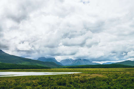 Dramatic landscape with mountain lake and forest on hills in sunlight and snowy mountains in low clouds in changeable weather. High mountains in cloudy sky and lake near green grasses and forest hillsの写真素材
