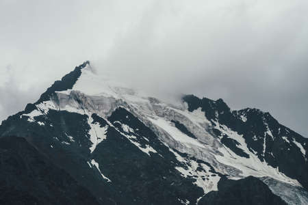 Minimalist monochrome atmospheric mountains landscape with big snowy mountain top in low clouds. Awesome minimal scenery with glacier on rocks. Blue white high mountain pinnacle with snow in clouds.の写真素材