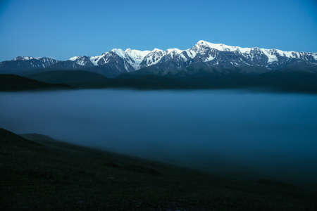 Atmospheric mountains landscape with dense fog and great snow mountain top under twilight sky. Alpine scenery with big snowy mountains over thick fog in night. High snow pinnacle above clouds in dusk.の写真素材