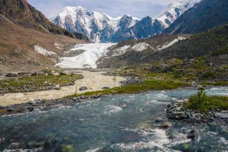 Sunny alpine landscape with confluence of two various mountain rivers on background of snowy mountain with glacier. Beautiful clear creek flows into dirty river. Confluence of two different rivers.の写真素材