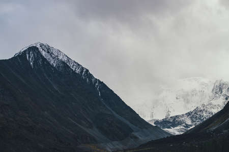 Dark atmospheric landscape with high black mountain top and snow-covered mountain wall in low clouds. Dramatic foggy mountain landscape with peaked top and snowy mountains in fog in overcast weather.の写真素材