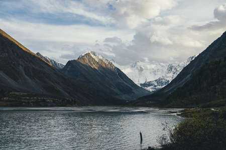 Awesome alpine landscape with mountain lake in highland valley and snow-covered mountain wall and rocks in golden sunlight under cloudy sky. Atmospheric scenery with pointy peaks and snowy mountains.の写真素材