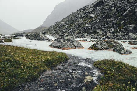 Atmospheric landscape with clear water stream flows into mountain river among moraines in rainy weather. Bleak scenery with milky river among rocks. Clear water stream flows into milk mountain river.の写真素材