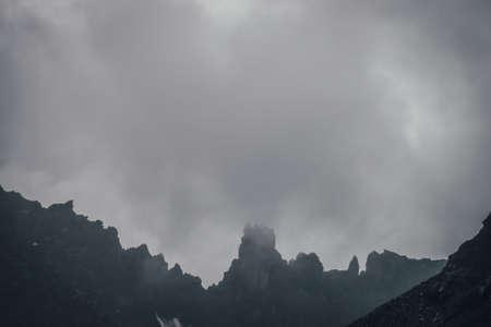 Dark atmospheric mountain landscape with black pointy rocky peak in gray cloudy sky. Lead gray low...