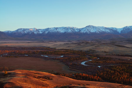 Awesome autumn landscape with mountain river in forest valley and great snow-covered mountain range...