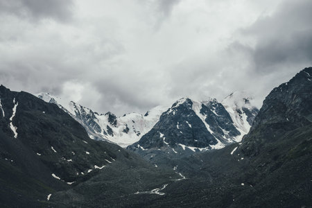 Dark atmospheric mountain landscape with glacier on black rocks in gray cloudy sky. Snowy mountains in low clouds in rainy weather. Gloomy mountain landscape with black rocky mountains with snow.の写真素材