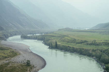 Beautiful misty mountain landscape with wide mountain river. Dark green gloomy scenery with big mountain river in mist. Dark atmospheric view to great river among big mountains in rainy weather.の写真素材