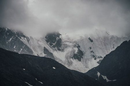 Dark atmospheric mountain landscape with glacier on black rocks in lead gray cloudy sky. Snowy mountains in low clouds in rainy weather. Gloomy landscape with black rocky mountains with snow in fog.の写真素材