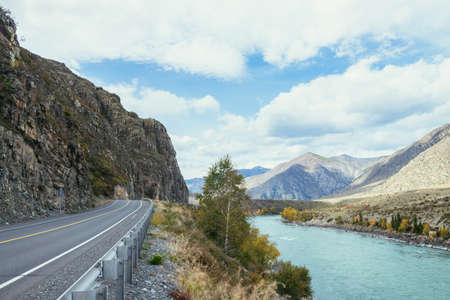 Vivid autumn landscape with mountain highway along big mountain river in sunshine. Bright alpine scenery with wide turquoise river and mountain road in autumn colors. Highway in mountains in fall timeの写真素材