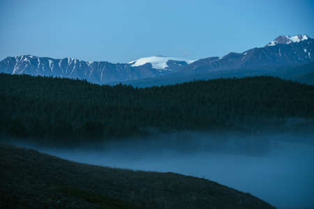Atmospheric mountains landscape with dense fog and great snow mountain top under twilight sky. Alpine scenery with big snowy mountains over thick fog in night. High snow pinnacle above clouds in dusk.の写真素材