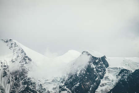 Atmospheric mountains landscape with big snowy mountain top with snow cornices in low clouds. Awesome minimal scenery with snow-white high pinnacle in overcast weather. Giant mountain wall in clouds.の写真素材
