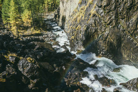 Atmospheric autumn landscape with rainbow above turbulent mountain river among rocks near rocky wall in sunshine. Beautiful alpine scenery with powerful mountain river and autumn forest in sunlight.の写真素材