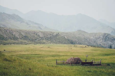 Beautiful foggy mountain landscape with scarecrow on wooden fence around haystack on background of mountains and rocks silhouettes in fog. Stack of hay is surrounded by wood fence in vintage tones.の写真素材