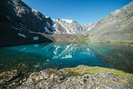 Snowy mountain reflected in clear water of glacial lake. Beautiful sunny landscape with glacier reflection in water surface of mountain lake under clear sky. Snow on rock reflected in mountain lake.の写真素材