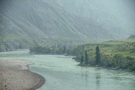 Beautiful misty mountain landscape with wide mountain river. Dark green gloomy scenery with big mountain river in mist. Dark atmospheric view to great river among big mountains in rainy weather.の写真素材