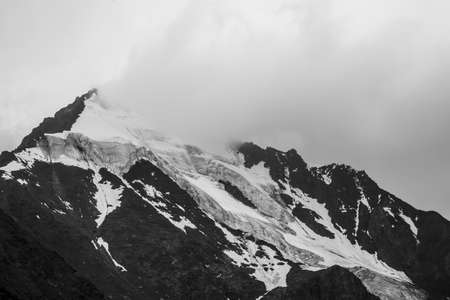 Monochrome atmospheric mountains landscape with big snowy mountain top in low clouds. Awesome minimal scenery with white glacier on black rocks. High mountain pinnacle with snow in clouds in grayscaleの写真素材
