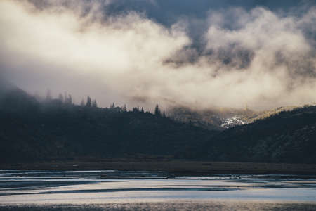 Mountain landscape with shiny lake and silhouettes of coniferous trees on dark rocks in low cloud in...