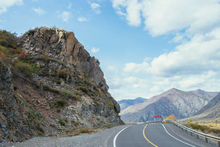Colorful autumn landscape with mountain highway along rocks in sunshine. Bright alpine scenery with mountain road in autumn colors. Highway in mountains in fall time. Road along beautiful rocky wall.の写真素材