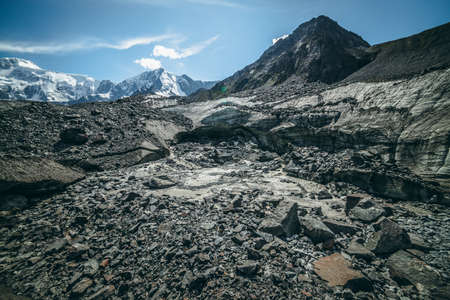 Scenic highlands landscape with big cracked glacier with scratches among moraines on background of high snowy mountain peak and rock top. Awesome scenery with large glacier and great snowy mountain.の写真素材