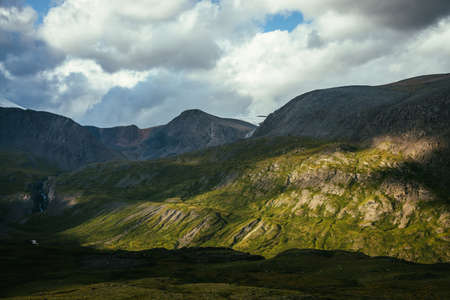 Dramatic alpine scenery with high mountain wall in sunlight and in shadow under cloudy sky. Scenic mountain landscape with changeable weather in highlands. Sunny and overcast. Sunlight and shadow.の写真素材