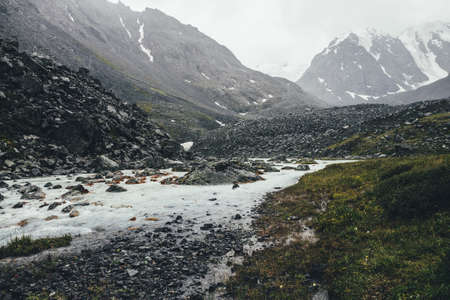 Atmospheric landscape with mountain creek among moraines in rainy weather. Bleak scenery with milky river from snowy mountains. Stones with moss and lichen in water stream. Mountain river among rocks.の写真素材