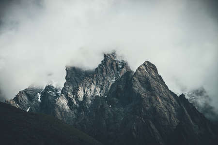 Dark atmospheric surreal landscape with dark rocky mountain top in low clouds in gray cloudy sky. Gray low cloud on high pinnacle. High black rock with snow in low clouds. Surrealist gloomy mountains.の写真素材