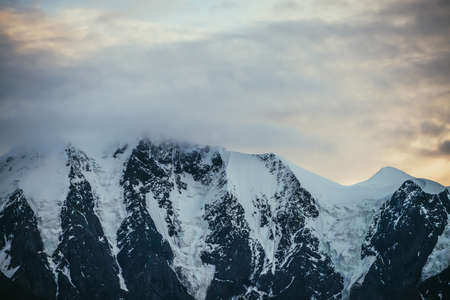 Minimal alpine landscape with snowy mountain top among low clouds on background of sunset or sunrise cloudy sky of illuminating color. Atmospheric scenery with snow-white mountain ridge in dawn sky.の写真素材