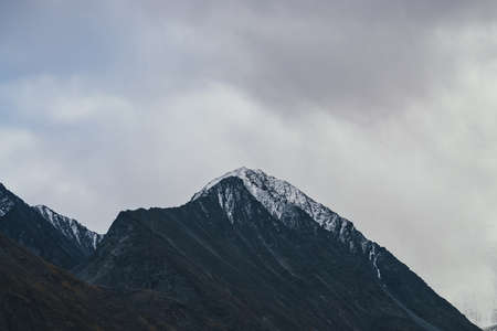 Dark atmospheric landscape with high black rocky mountain top with snow in cloudy sky. Dramatic mountain landscape with snow peaked top in haze in overcast weather. Gloomy view to awesome rocks in fogの写真素材