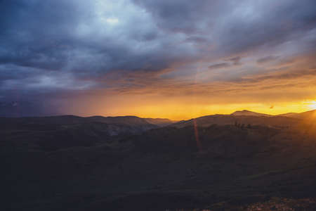 Atmospheric landscape with silhouettes of mountains with trees on background of dawn sky with vivid orange sunlight and sun rays. Colorful nature scenery with sunset or sunrise of illuminating color.の写真素材